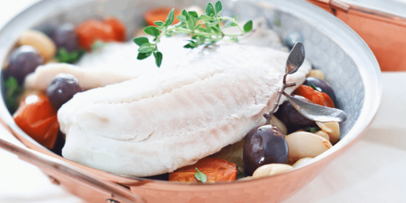 Filet de poisson blanc cuit dans une poêle en cuivre, entouré de tomates cerises, d'olives noires, de gousses d'ail et d'herbes.