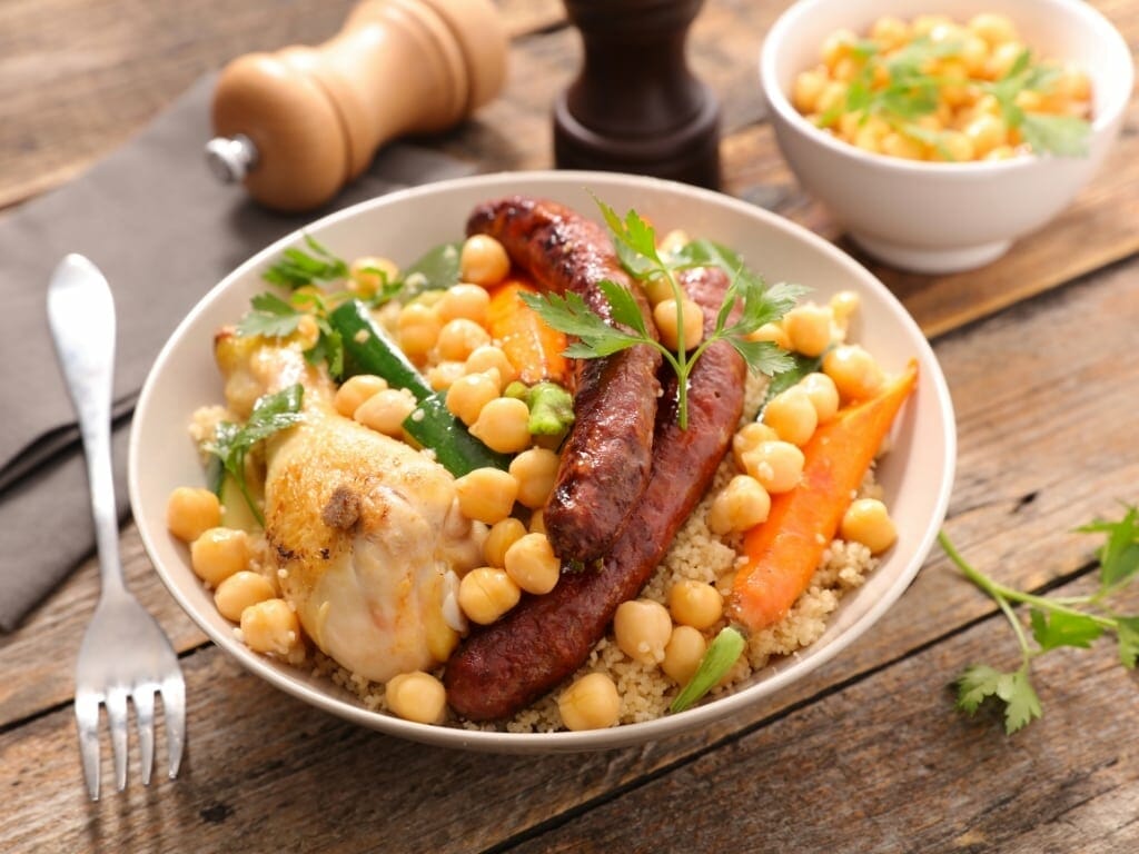 Un bol de couscous avec un pilon de poulet, deux saucisses, des pois chiches, des courgettes et des carottes. Un petit bol de pois chiches et un moulin à sel et à poivre sont visibles à l'arrière-plan.
