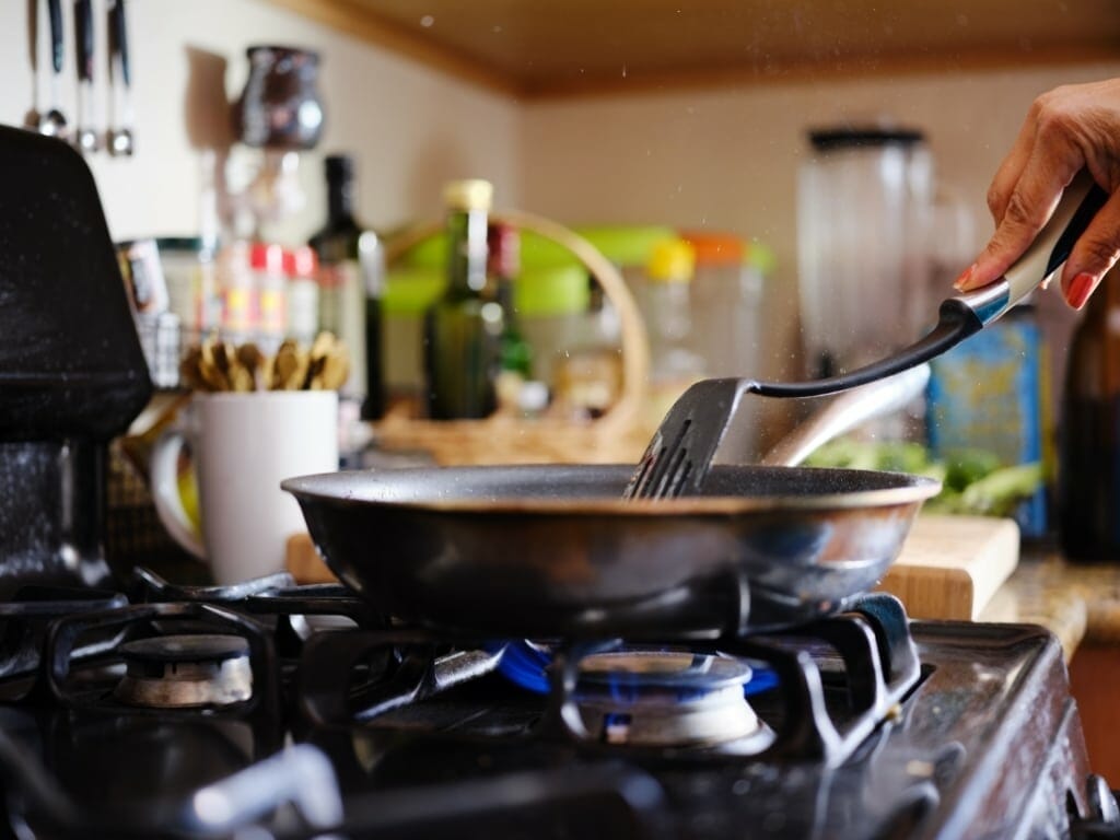 Une personne utilise une spatule pour cuisiner dans une poêle sur une cuisinière. Le plan de travail, en arrière-plan, est occupé par divers ustensiles et récipients.