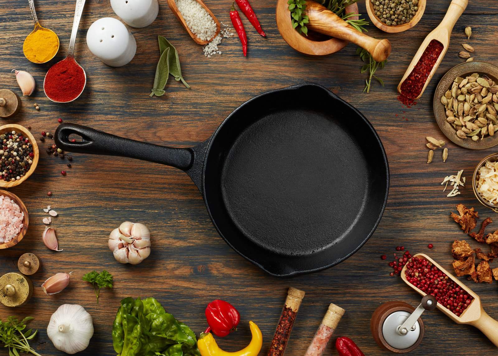 Une poêle en fonte entourée de diverses épices, herbes, ail, poivrons et légumes sur une surface en bois.