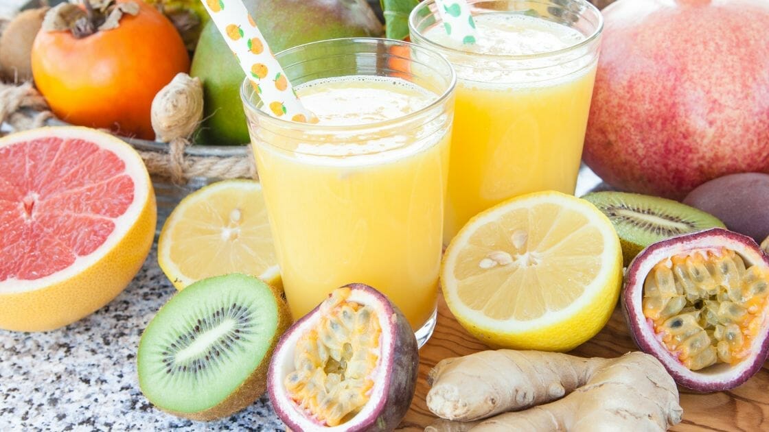 Deux verres de jus d'orange avec des pailles entourés de divers fruits, dont du pamplemousse, du kiwi, du citron, du fruit de la passion, du gingembre et une grenade sur une surface mouchetée.