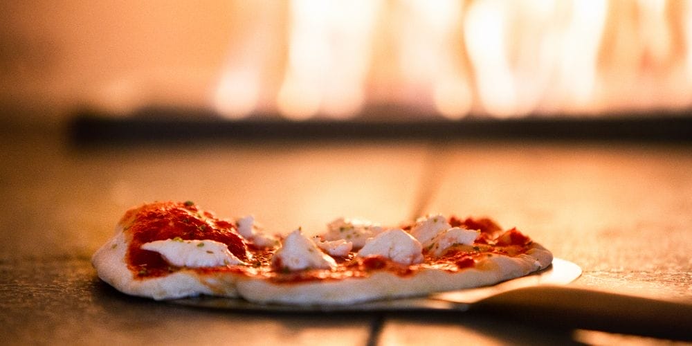 Gros plan d'une pizza au fromage et à la tomate placée sur une pelle devant un four à bois allumé.