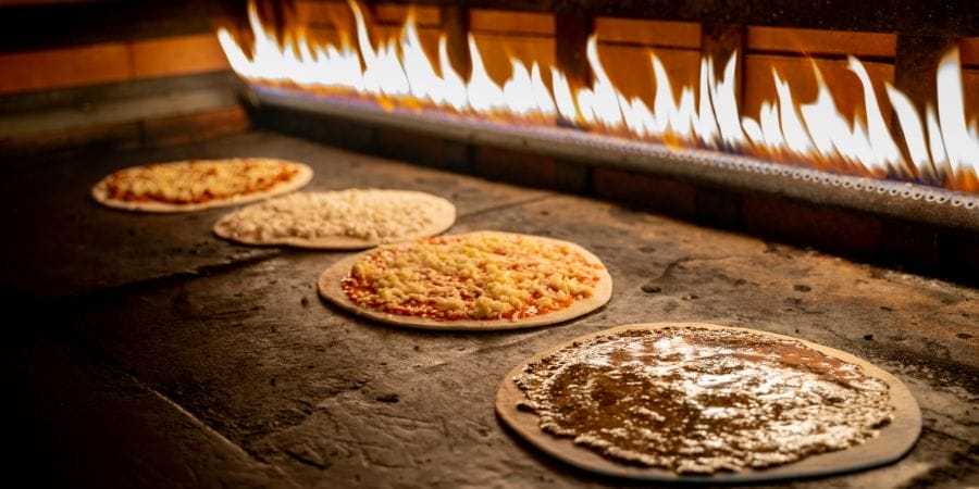 Quatre pizzas cuisent sur une surface en pierre dans un four à bois, avec des flammes visibles en arrière-plan.