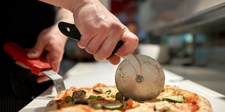 Une personne coupe une pizza garnie de légumes avec un coupe-pizza, en tenant la tarte fermement avec des pinces.